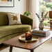 A lit Tortoise Glass candle sits on a round brass tray on a wooden coffee table in a styled living room with an olive green sofa and a window view.