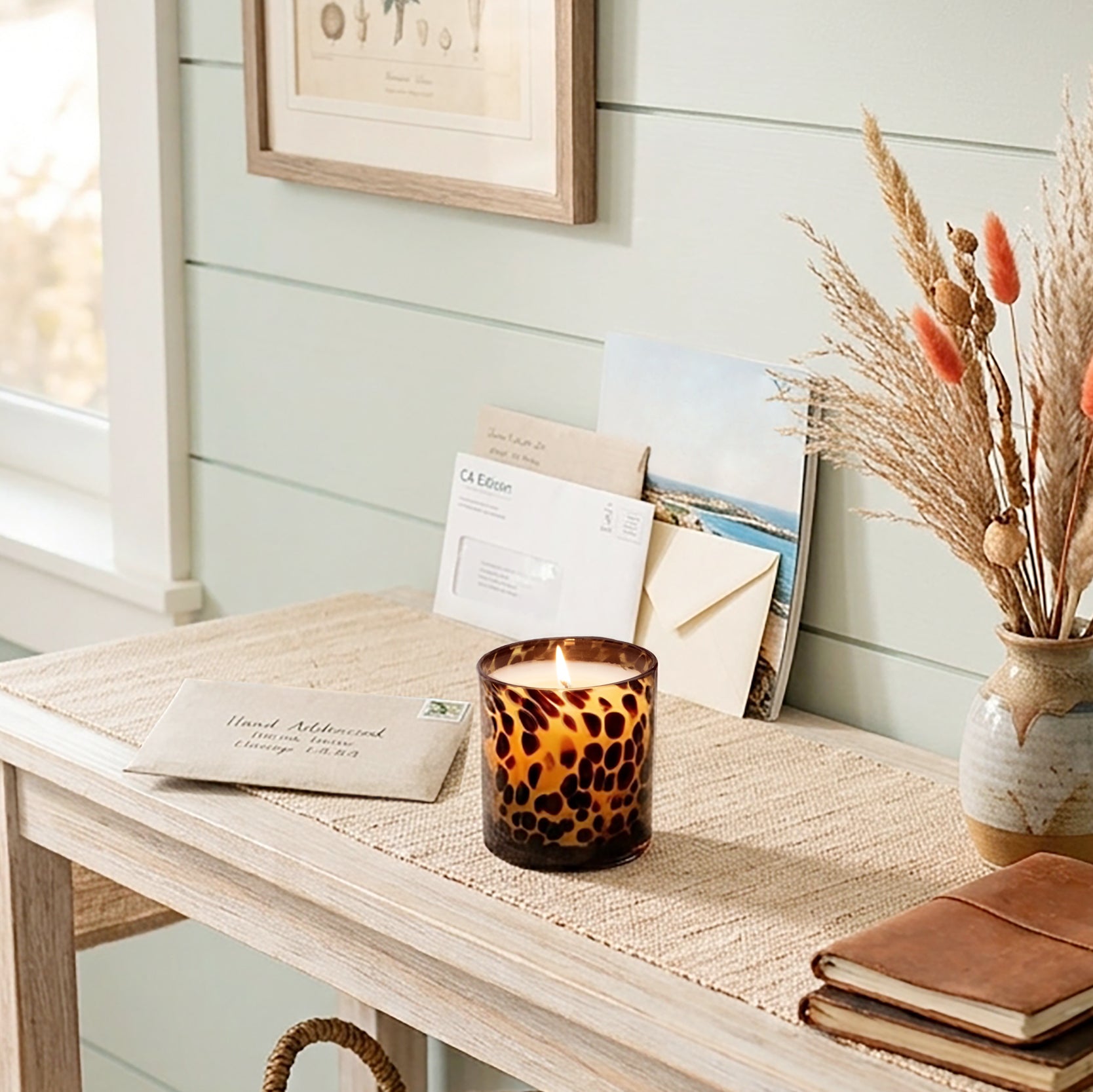 A lit Tortoise Glass candle sits on a light wood console table in a styled room setting with a woven runner, dried grasses in a ceramic vase, and a notebook.