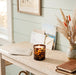 A lit Tortoise Glass candle sits on a light wood console table in a styled room setting with a woven runner, dried grasses in a ceramic vase, and a notebook.