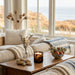 A cozy living room scene with a wooden coffee table holding a lit Tortoise Glass candle, next to a small glass vase with dried stems, and a bowl of fruit. A cream sofa with a beige throw blanket and a blue-striped pillow is in front of large windows overlooking a beach and dunes.