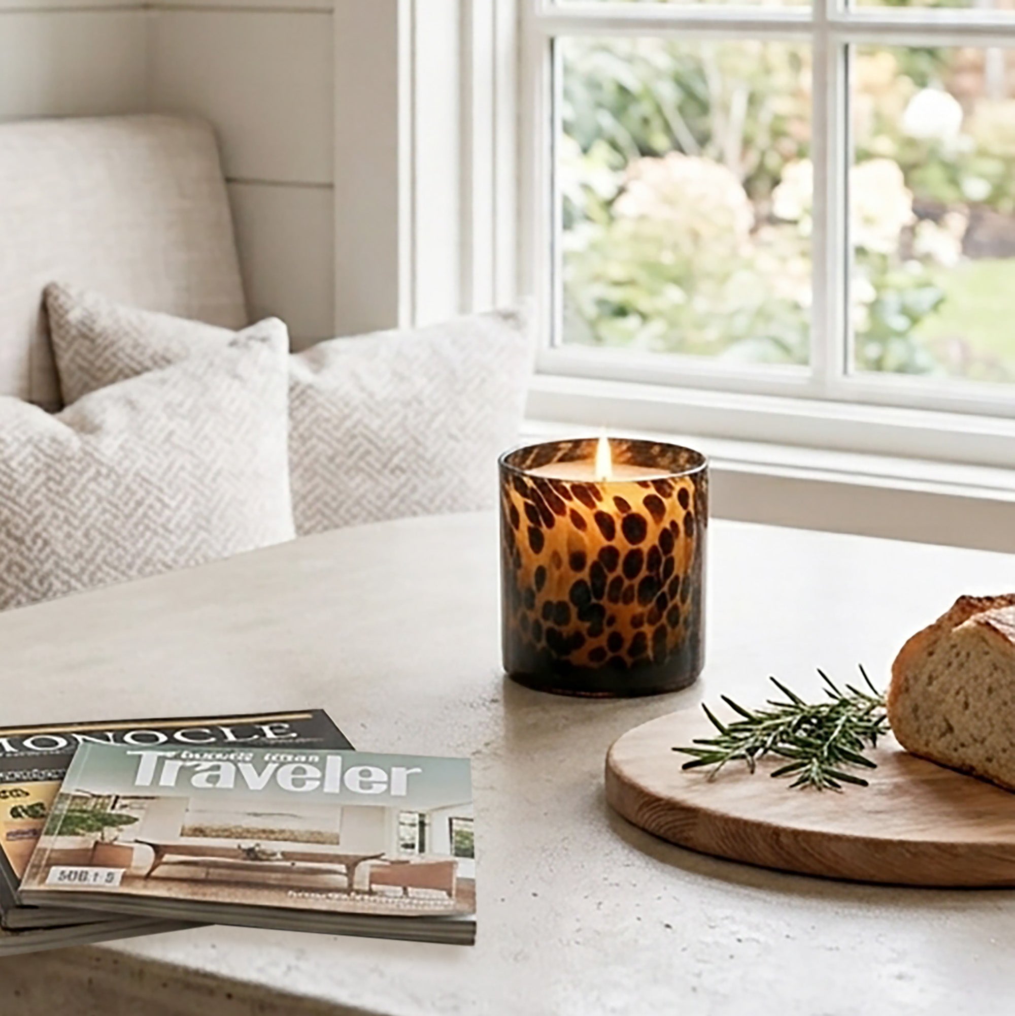 Sitting on a light stone table in a cozy room setting, with a window, pillows, a magazine, and a loaf of bread on a wooden board nearby, is a lit Tortoise glass candle.
