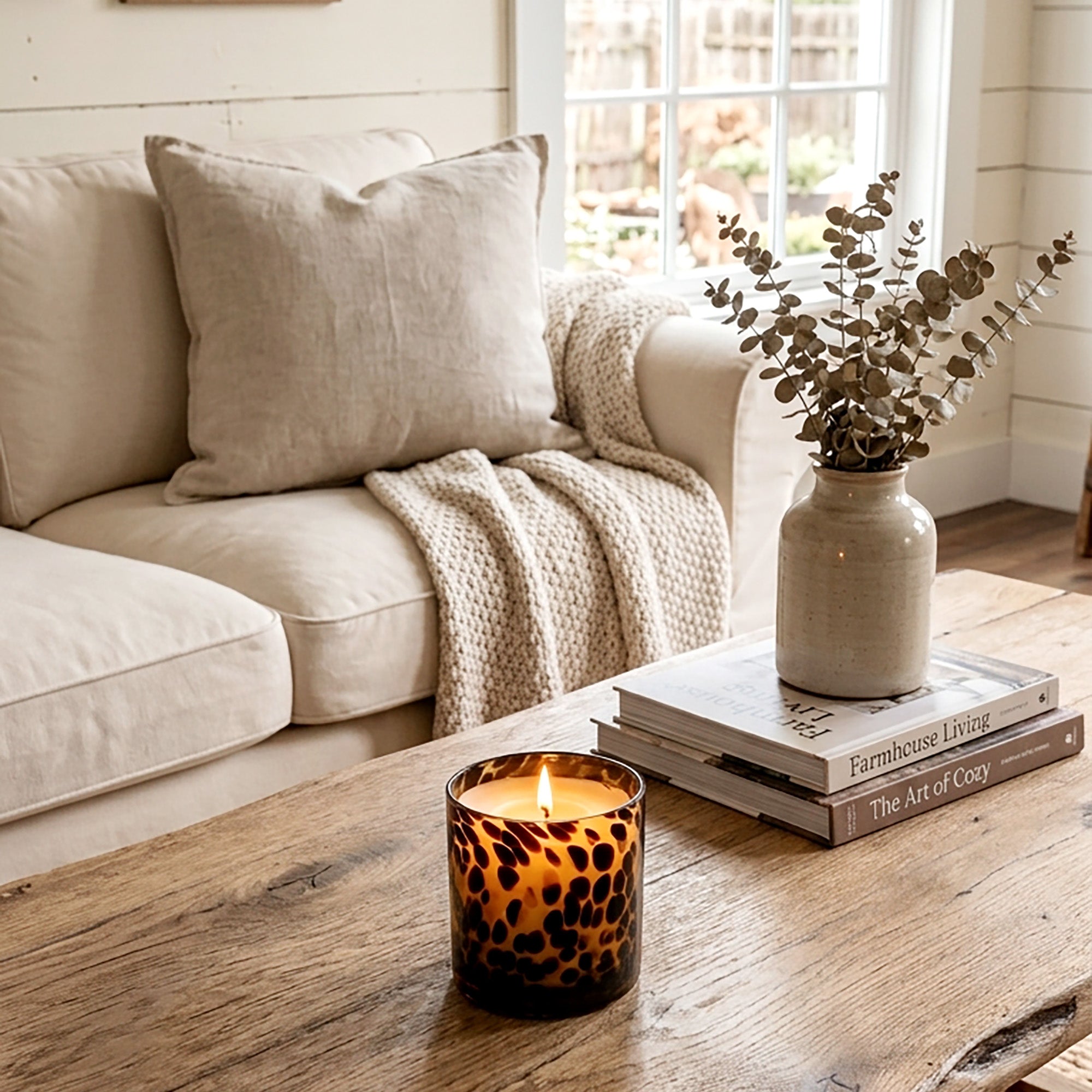 Sitting on a rustic wooden table in a cozy living room setting, next to a stack of books and a ceramic vase with eucalyptus, is a lit Tortoise Glass candle.  A beige sofa is in the background.