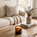 Sitting on a rustic wooden table in a cozy living room setting, next to a stack of books and a ceramic vase with eucalyptus, is a lit Tortoise Glass candle.  A beige sofa is in the background.