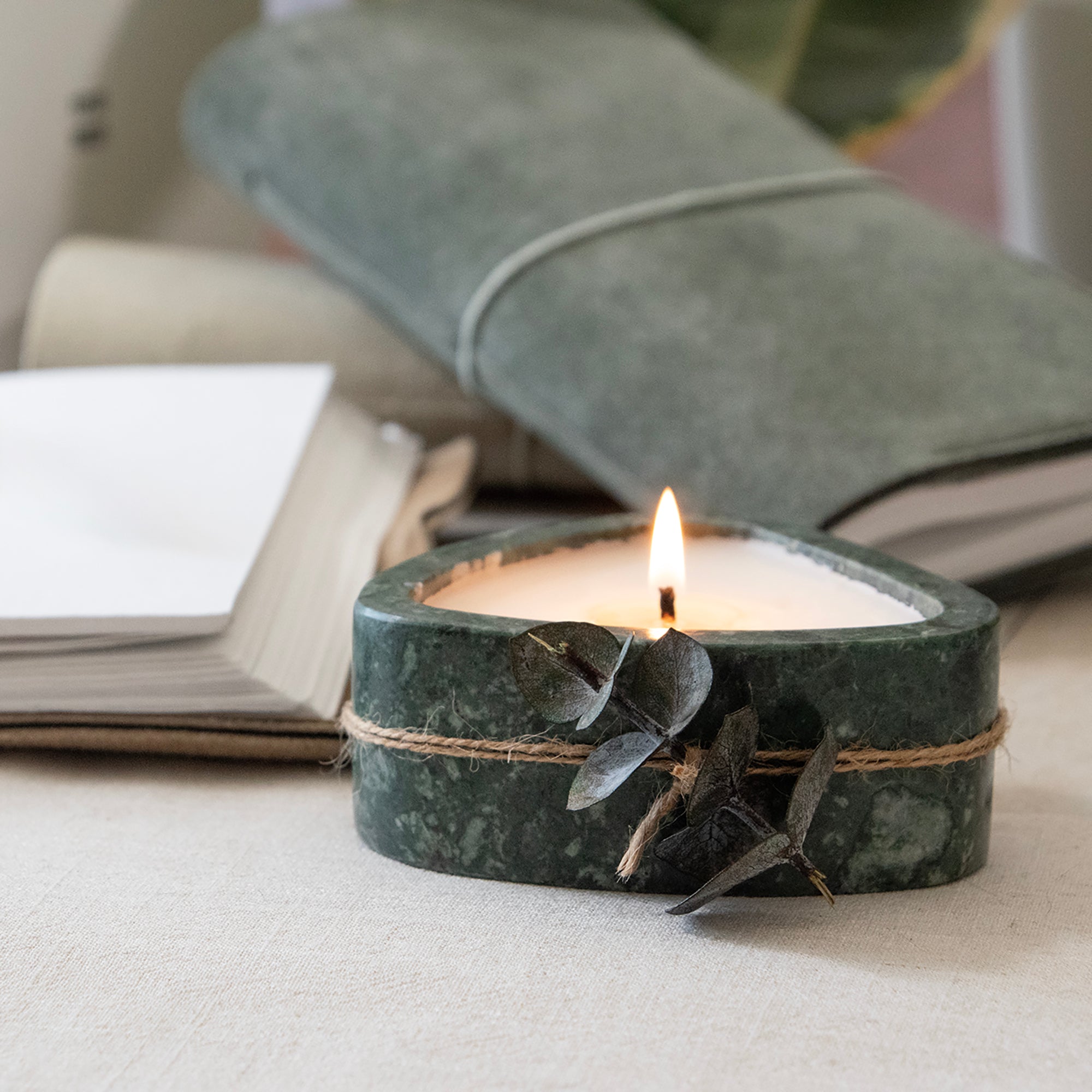 Candle in a green marble holder with leaves on a light surface, with books and a pillow in the background.