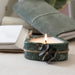 Candle in a green marble holder with leaves on a light surface, with books and a pillow in the background.
