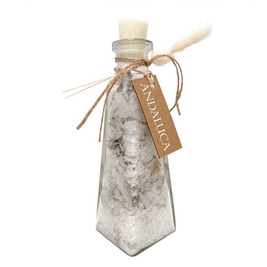 A clear glass bottle with a pointed bottom, filled with white salt crystals, sealed with a cream-colored wax or beeswax plug. The bottle is decorated with a rustic brown twine bow, a small beige feather, and a brown kraft paper tag that reads 'ANDAUCA'.