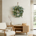 A green eucalyptus wreath named Willow and Silver Dollar Eucalyptus Wreath is hanging on a textured beige wall above a light wood console table in a modern, minimalist living room setting.