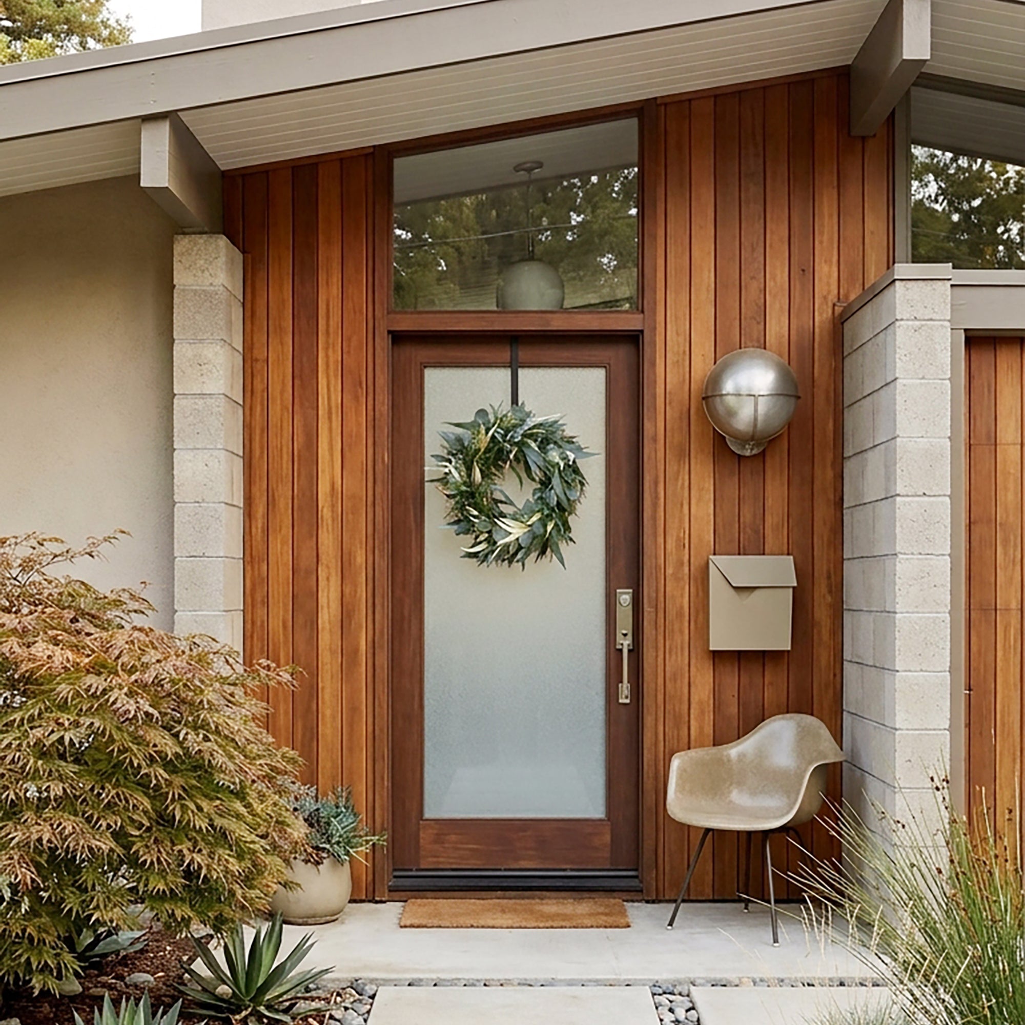 A modern front door with warm wood siding, a frosted glass door panel, a green wreath named Willow and Silver Dollar Eucalyptus Wreath is hanging on the door. A beige chair, and a metal mailbox are shown in an outdoor residential setting.