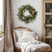 Winter Sage Haoliday wreath hung on a wall above a patterned chair with pillows and a blanket.