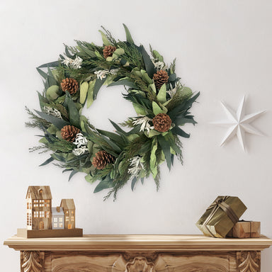 Winter Sage Haoliday wreath hung  on a white wall above a wooden mantel with decorative items.