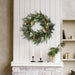Winter Sage Haoliday wreath hung  on a white wall above a fireplace with candles and decorative items.