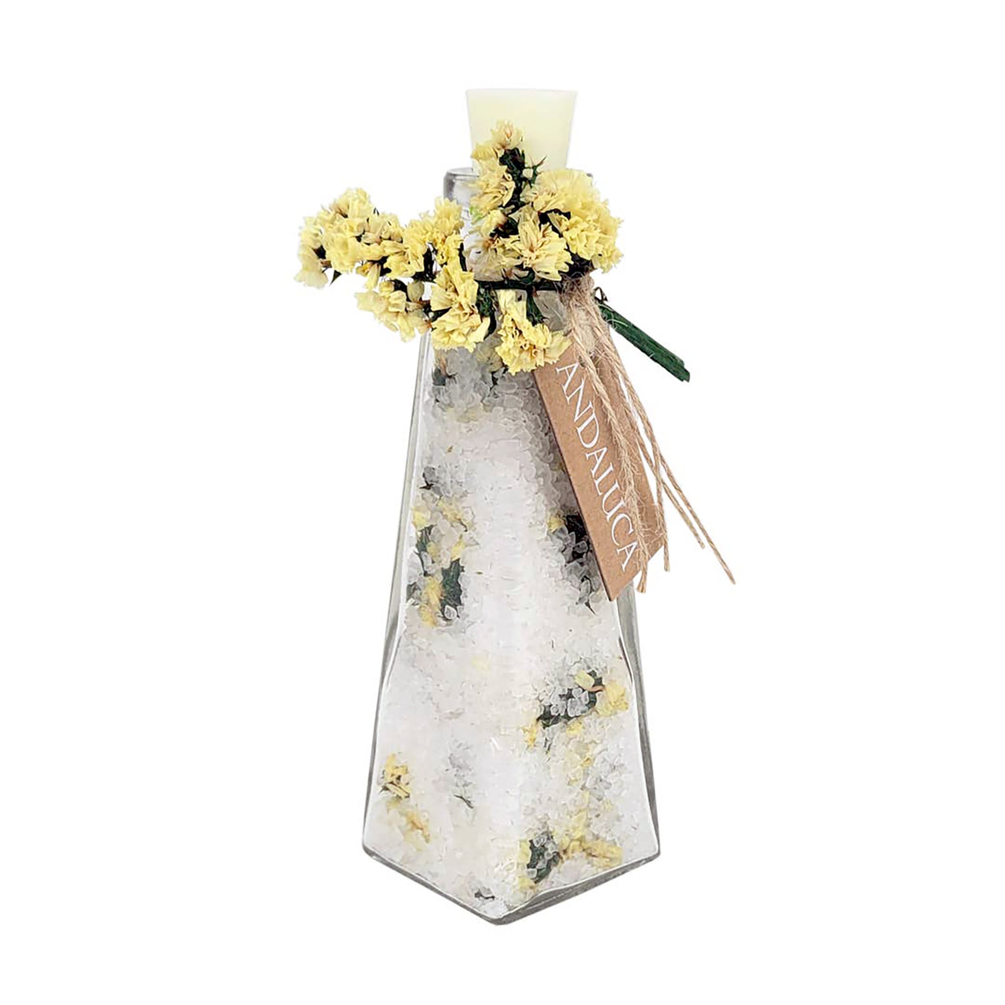 A clear glass bottle with a faceted hexagonal shape, filled with white bath salts and decorated with yellow dried flowers and green leaves. A light brown paper tag tied with a ribbon is attached to the neck, featuring the brand name 'ANDALUCIA'.